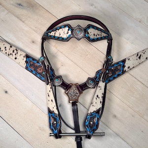 May include: A horse bridle and breast collar set featuring white cowhide with black spots, accented by bright blue floral tooling and silver-toned conchos with turquoise-colored centers. The leather components are dark brown with metal hardware.