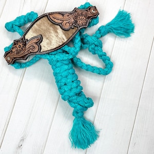 May include: A turquoise rope halter with a tooled leather browband. The browband features a light brown hide center and intricate floral tooling. The halter has a braided design and a tassel at the end.