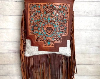 May include: A brown leather crossbody bag with a turquoise floral design. The bag features a long fringe and a white and gray cowhide panel. The bag has a long adjustable strap.