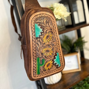 Puede incluir: Una bolsa de bandolera de cuero marrón con un diseño estampado que presenta girasoles, un cactus y detalles en turquesa.