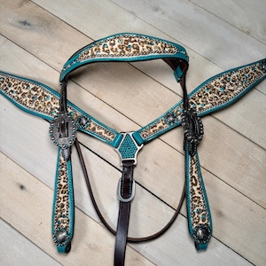 May include: A turquoise and brown leather horse bridle set with a leopard print design. The set includes a headstall, breast collar, and reins, all adorned with turquoise accents and silver-tone hardware. The bridle is designed for equestrian use.