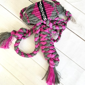 May include: A braided rope halter with pink and gray strands. The halter has a decorative woven panel with black and white stripes. The halter has tassels on the ends.