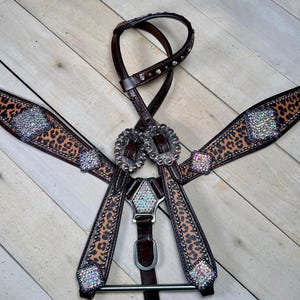 Puede incluir: Un petral de caballo de cuero marrón y beige con un diseño de estampado de leopardo. El petral está adornado con hebillas plateadas y brillantes pedrería iridiscente. La brida también es de cuero marrón con tachuelas plateadas.