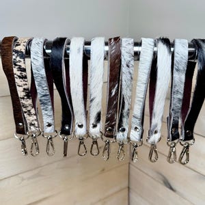 May include: A collection of leather wrist straps with various cowhide patterns, including black and white, brown and white, and solid black. Each strap has a silver-colored clasp for attaching to a bag or keys.
