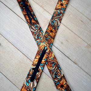Puede incluir: Dos correas largas y estrechas con un vibrante patrón floral y de volutas en tonos naranja, marrón y turquesa brillante. Cada correa incluye una fina franja vertical negra en el centro, creando una disposición en forma de cruz.