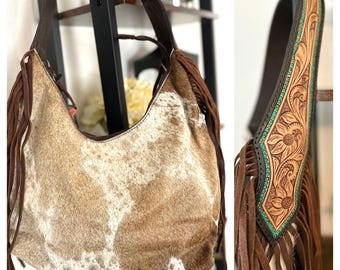 May include: A brown and white cowhide handbag with fringe detailing. The bag has a brown leather strap with a tooled design and turquoise accents.