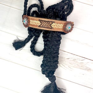May include: Black horse halter with a decorative leather browband. The browband features a woven design with turquoise and bronze accents. The halter is made of braided rope and includes a tassel.