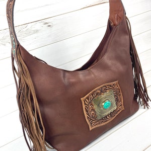 Puede incluir: Bolso hobo de cuero marrón con detalles de flecos y un diseño de cuero repujado con una piedra turquesa.