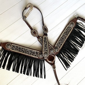 May include: A brown leather horse breast collar with decorative silver-toned metal accents and black fringe. The breast collar features intricate detailing and is designed for equestrian use.
