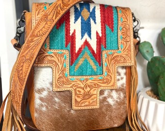 May include: A brown leather crossbody bag with a fringe detail. The bag has a turquoise, red, white, and blue geometric pattern on the front. The bag is made of cowhide and has a tooled leather design.