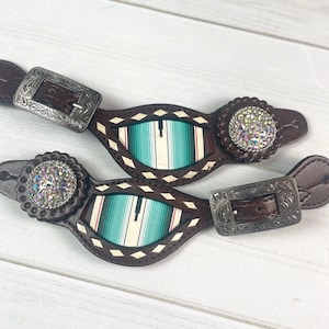 May include: A pair of brown leather boot straps with turquoise and red patterned fabric inserts. Each strap has a silver buckle and a round, clear rhinestone embellishment.