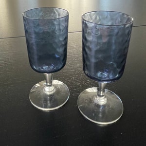 May include: Two clear glass wine glasses with a textured, blue-tinted surface. The glasses have a stem and a round base.