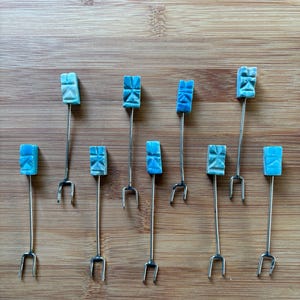 May include: A collection of ten small, decorative forks. Each fork has a silver-colored stem and a two-pronged end. The top of each fork features a square, turquoise-colored decorative element with carved details. The forks are arranged on a wooden surface.