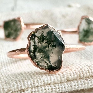 Peut inclure: Trois bagues ornées de pierres d'agate mousse brutes et irrégulières avec des inclusions dendritiques vert foncé. Chaque pierre est montée sur un anneau fin en cuivre poli. Une bague est au premier plan, deux autres sont en arrière-plan.
