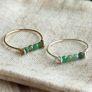May include: Two delicate rings with a row of green gemstone beads. The ring on the left is gold and the ring on the right is silver.