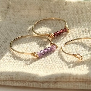 Op de afbeelding: Drie gouden ringen met verschillende gekleurde edelstenen. De ringen liggen op een beige stoffen oppervlak. De edelstenen zijn amethyst, granaat en helder.