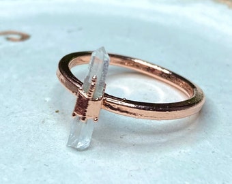 Könnte beinhalten: Ein Ring aus Roségold mit einer klaren Quarzkristallspitze. Der Ring liegt auf einer weißen Keramikplatte.