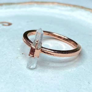 May include: A rose gold ring featuring a clear quartz crystal point. The ring is displayed on a white ceramic dish.