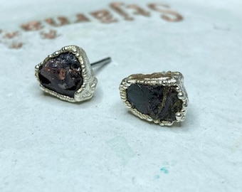 May include: A pair of silver stud earrings with a dark red, rough-cut gemstone set in each. The earrings have a textured silver border.