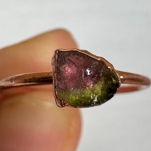 May include: A ring featuring a raw, uncut bicolour tourmaline gemstone set on a thin, polished copper band. The stone displays a distinct colour gradient, transitioning from a deep pinkish-red at the top to a vibrant olive green at the base.