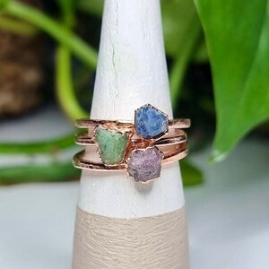 Puede incluir: Tres anillos con piedras preciosas en bruto engastadas en oro rosa. Los anillos presentan un zafiro azul, una esmeralda verde y una turmalina rosa.