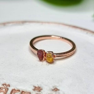 Könnte beinhalten: Roségoldener Ring mit zwei rohen Edelsteinen, einem rosa und einem gelben. Der Ring hat ein dünnes Band, und die Edelsteine sind eng beieinander gesetzt. Der Ring wird auf einer weißen Oberfläche präsentiert.