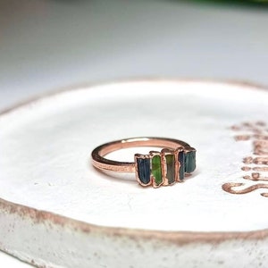 May include: A rose gold ring with a row of five green and blue gemstones. The ring is on a white ceramic dish.