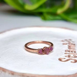 May include: A rose gold ring with three pink gemstones set in the band. The ring is sitting on a white ceramic dish.