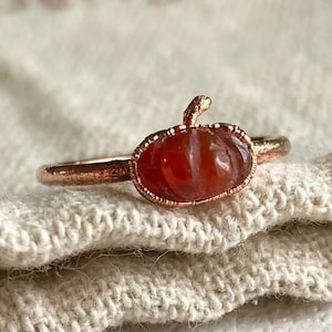May include: A rose gold ring featuring a small, carved, orange-red carnelian gemstone shaped like a pumpkin. The metal band has a textured, rustic finish, and the gemstone is set within a delicate, beaded metal frame that mimics the pumpkin's stem.