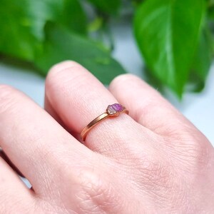 May include: A delicate gold ring with a small, pink, raw ruby gemstone set in the center.