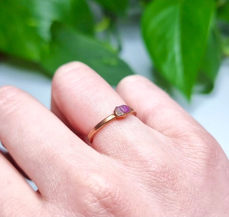 May include: A delicate gold ring with a small, pink, raw ruby gemstone set in the center.