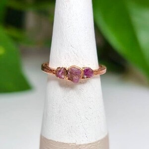 May include: A rose gold ring with three pink raw ruby stones set in the band.