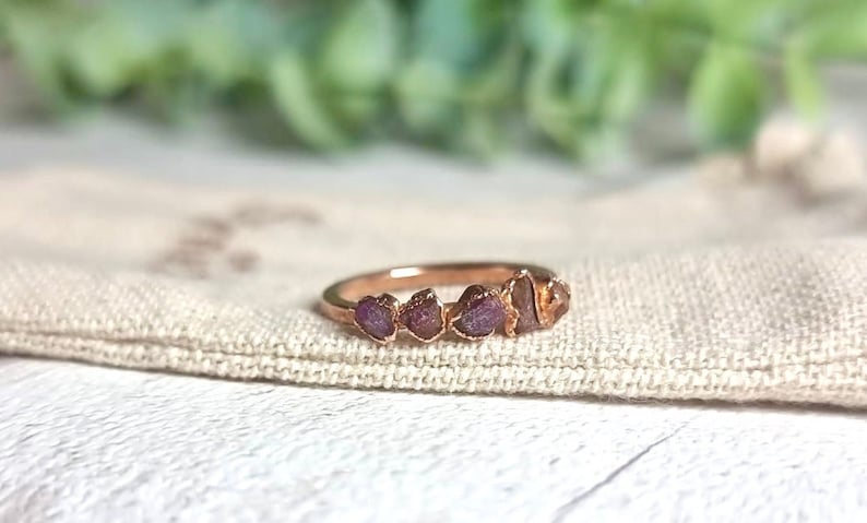 May include: A rose gold ring with five small, raw ruby stones set in a row. The ring is on a light brown fabric surface.