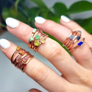 May include: A set of five rings with different colored gemstones wrapped around a copper band. The gemstones are green, yellow, orange, pink, and blue.