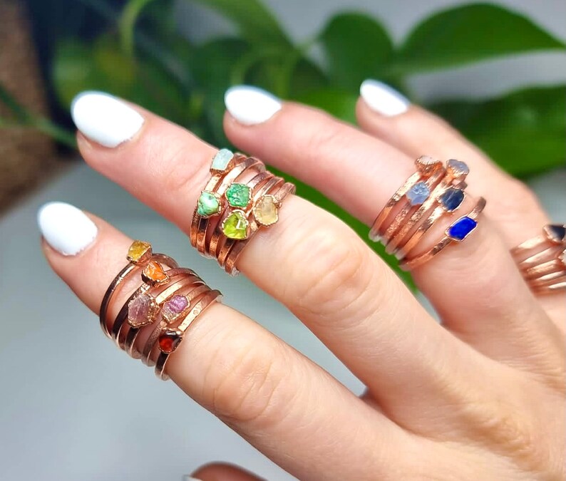 May include: A set of five rings with different colored gemstones wrapped around a copper band. The gemstones are green, yellow, orange, pink, and blue.