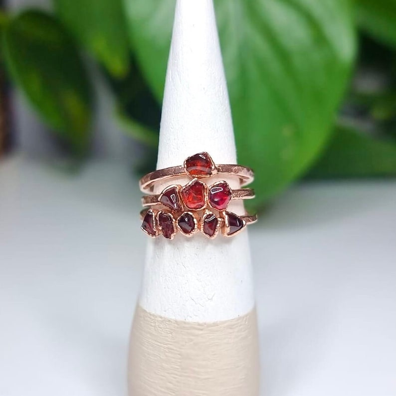 May include: A set of three rose gold rings with raw garnet stones. The rings are stacked on a white display stand.