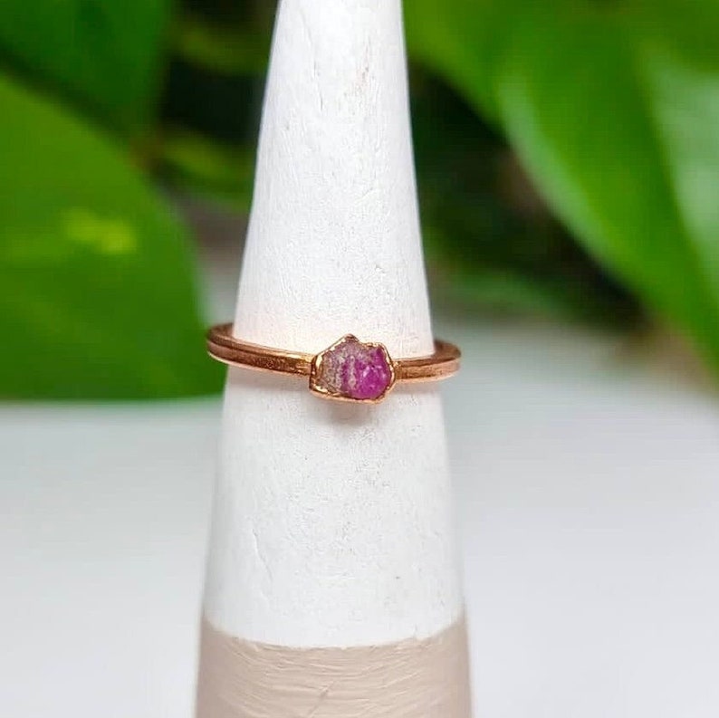 May include: A rose gold ring with a small, raw pink ruby gemstone set in the band.