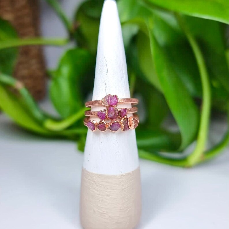 May include: Three rose gold rings with pink raw ruby stones set in the metal. The rings are stacked on a white and beige display stand.