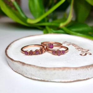 May include: Three rose gold rings with pink gemstone accents. The rings are stacked on a white ceramic dish.