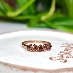 May include: A rose gold ring with five small, irregular, dark red gemstones set in a row. The ring is on a white ceramic dish with the word "Stay Wild" written on it.
