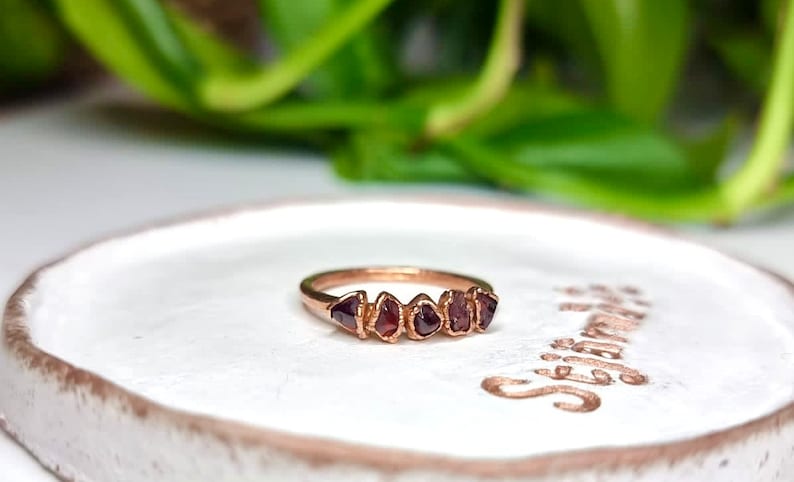 May include: A rose gold ring with five small, irregular, dark red gemstones set in a row. The ring is on a white ceramic dish with the word "Stay Wild" written on it.