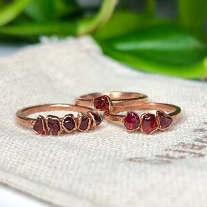 May include: Two gold rings with a row of small, red garnet stones set in the band. The rings are on a white fabric surface.
