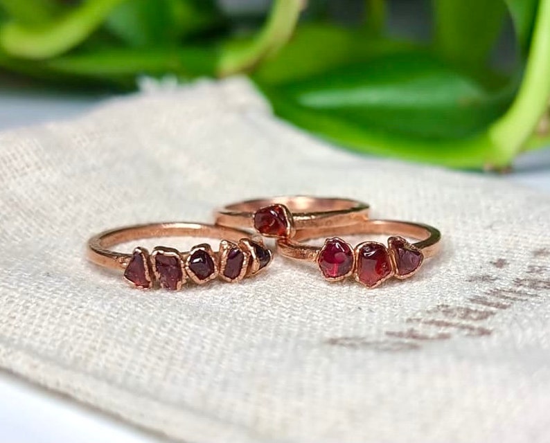 May include: Two gold rings with a row of small, red garnet stones set in the band. The rings are on a white fabric surface.