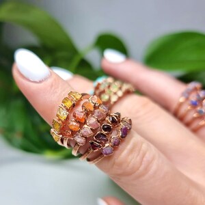 May include: A set of five gold rings with different colored gemstones. The rings are stacked on a finger and feature a variety of colors, including yellow, orange, pink, red, and purple.