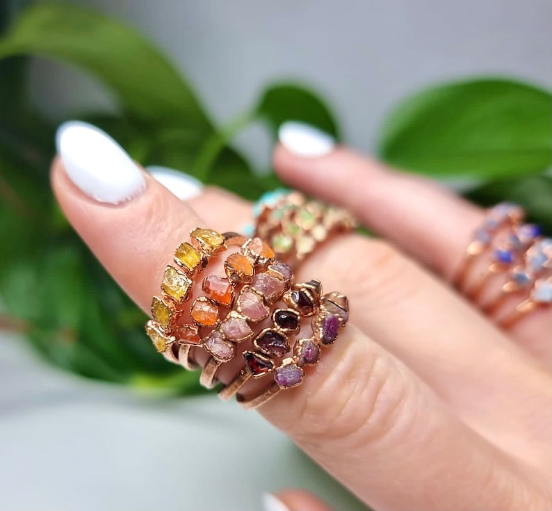 May include: A set of five gold rings with different colored gemstones. The rings are stacked on a finger and feature a variety of colors, including yellow, orange, pink, red, and purple.