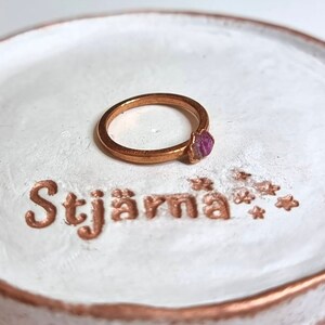 May include: A gold ring with a purple gemstone set on a white ceramic ring dish with the word "Stj&auml;rna" written in gold lettering.