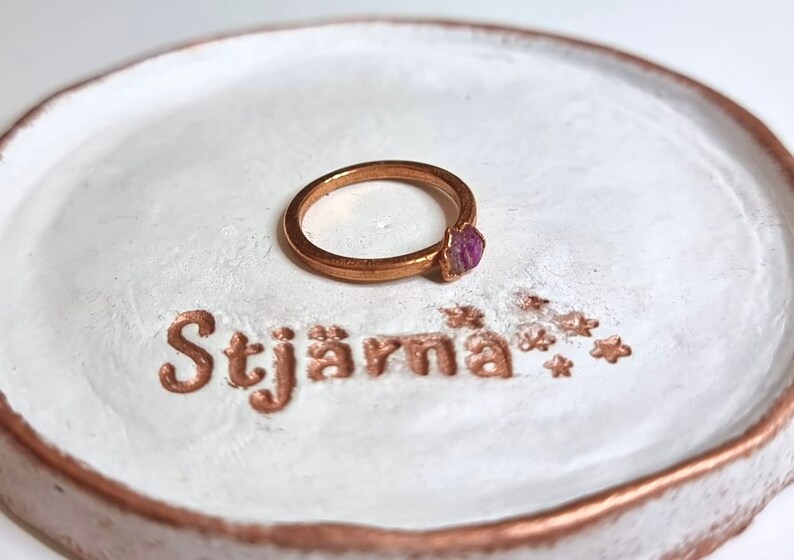 May include: A gold ring with a purple gemstone set on a white ceramic ring dish with the word "Stj&auml;rna" written in gold lettering.