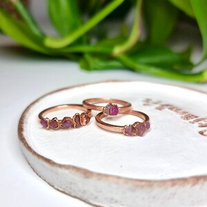 May include: Three rose gold rings with pink gemstone accents. The rings are on a white ceramic dish.