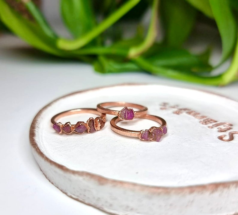 May include: Three rose gold rings with pink gemstone accents. The rings are on a white ceramic dish.