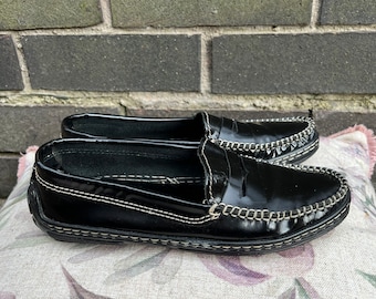 Könnte beinhalten: Ein Paar schwarze Leder-Slipper mit weißen Nähten. Die Slipper haben eine spitze Zehenpartie und einen niedrigen Absatz.